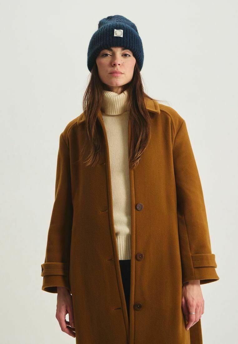 Manteau en laine brun avec un col à revers et des boutons, porté par-dessus un col roulé crème. Bonnet tricoté bleu foncé avec une étiquette à l'avant.