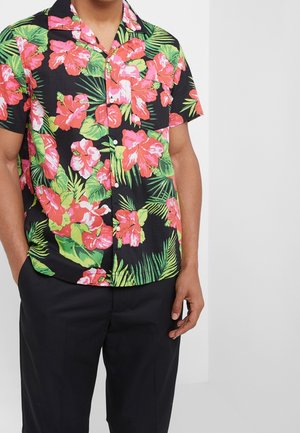 Homme portant une chemise noire à manches courtes avec un grand imprimé floral rose et vert, associée à un pantalon noir.
