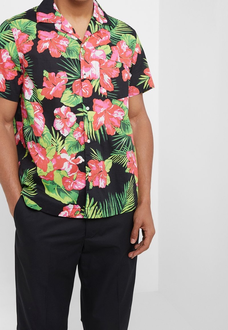 Homme portant une chemise noire à manches courtes avec un grand imprimé floral rose et vert, associée à un pantalon noir.