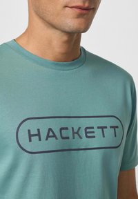Tyrkysové bavlněné tričko s tmavě námořnickým obdélníkovým logem "HACKETT". Kulatý výstřih, krátké rukávy a hladká textura.