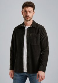 Camisa de pana oscura con botones en la parte delantera, mangas largas, bolsillo en el pecho y una textura suave, superpuesta sobre una camiseta blanca.