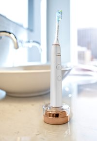 Philips Sonicare elektrische Zahnbürste mit blauen und gelben Borsten, die auf einem roségoldenen Sockel auf einer Badezimmertheke steht.