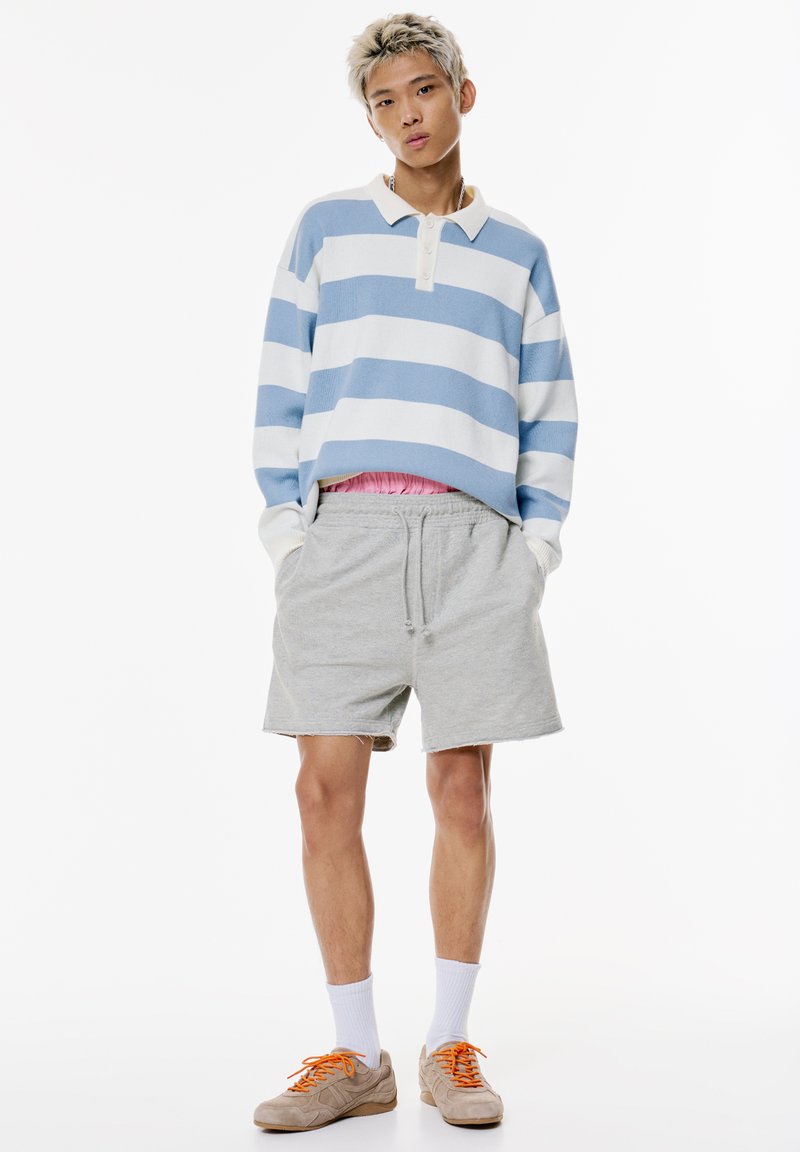 Blau-weißer gestreifter Langarmhemd mit Kragen, graue Shorts, weiße Socken und beige Turnschuhe mit orangefarbenen Schnürsenkeln.