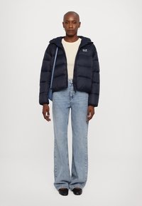 Mornarska puffer jakna s kapuco in z zadrgo, s potiskom na prsih, v kombinaciji z visokimi jeans hlačami v bledi barvi in črnimi čevlji.