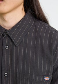 Fekete galléros ing függőleges bézs csíkokkal, fekete gombokkal, bal oldali mellzsebbel és Dickies logós címkével. Texturált anyag.