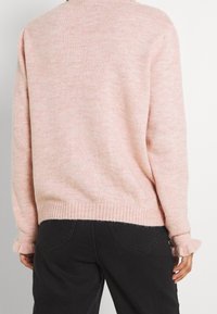 Osoba v světle růžovém pleteném svetru s žebrovaným lemem a manžetami, spárovaném s černými kalhotami s vysokým pasem, zobrazená zezadu.