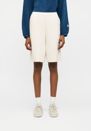 Άτομο με κρεμ βερμούδα μέχρι το γόνατο, λευκές κάλτσες, μπεζ αθλητικά παπούτσια και μπλε μπλούζα με μακριά μανίκια μπροστά σε απλό φωτεινό φόντο.