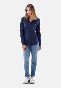 Donna in piedi con postura rilassata, che indossa una camicia blu navy con bottoni, jeans azzurri e sneakers bianche con strisce nere.