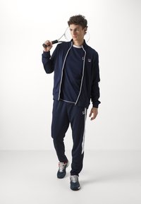 Marineblaues Sportanzug mit weißen Akzenten; Jacke mit Reißverschluss, passende Hose und sportliche Sneakers. Die Textur ist glatt, der Stoff wirkt leicht.