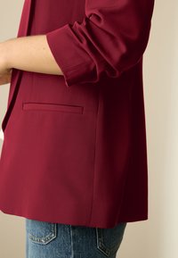 Sacou bordo cu design structurat, țesătură texturată și o singură buzunar la piept, asortat cu denim albastru.