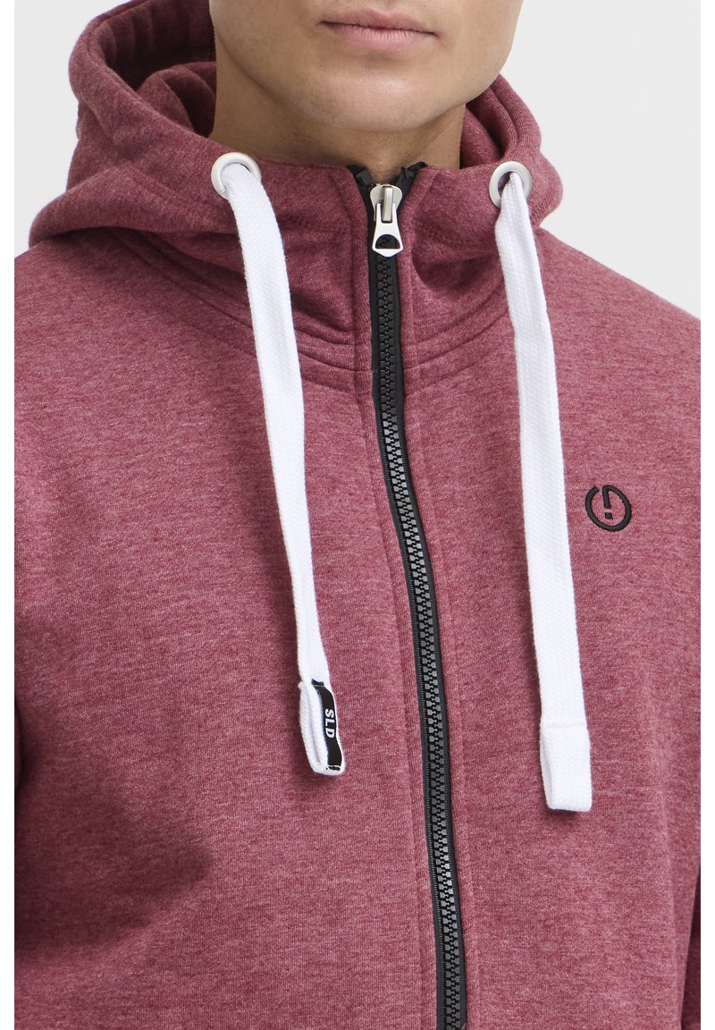 Rode sweatshirt met capuchon, voorzien van een zwarte rits, witte trekkoorden en een klein logo op de borst. Zacht, zichtbaar textuurstof.