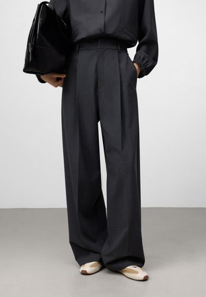 Wijdvallende, donkergrijze broek met een textuurafwerking, voorzien van een hoge taille en subtiele plooien. Gecombineerd met crèmekleurige en bruine schoenen.