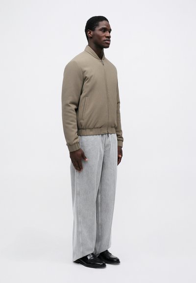 Χακί bomber jacket με ριμπ λάστιχα στους καρπούς και στο στρίφωμα, συνδυασμένο με φωτεινά γκρίζα wide-leg jeans και μαύρα παπούτσια, εκτεθειμένο σε ουδέτερο φόντο.