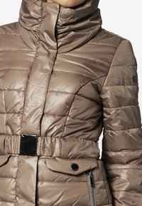 Kurtka puchowa z wysokim kołnierzem, pikowana, w jasnym odcieniu taupe. Posiada regulowany pas, błyszczące wykończenie oraz kieszeń na zamek błyskawiczny.