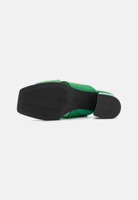 Chaussure en cuir vert avec un talon carré, semelle noire texturée, design à large lanière et finition en matériau doux. La vue inférieure montre le motif de la semelle.