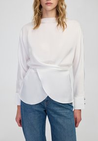 Vrouw in een witte blouse met lange mouwen en gelaagde voorkant, gecombineerd met blauwe jeans, gefotografeerd van halverwege de borst tot de heupen tegen een effen achtergrond.