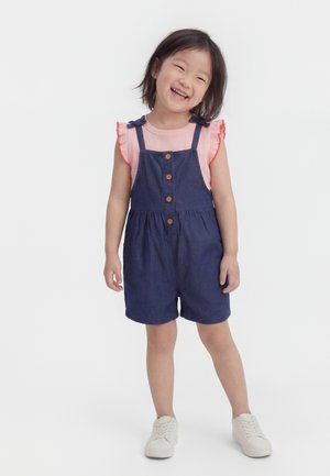 Smiling young girl with short dark hair wearing a pink ruffle-sleeve shirt under navy blue short overalls and white sneakers standing.