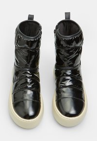 Botas de tobillo negras y brillantes con diseño acolchado, detalles de costura en la parte frontal, suela color beige claro y tirador trasero. Textura suave y parte superior acolchada.