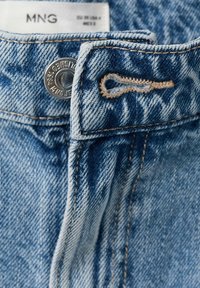 Detail svetlomodrých riflí z denimu s kovovým gombíkom, gombíkovou dierkou a bielou štítkou s označením značky MNG a veľkostnými údajmi.