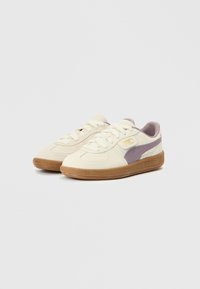 Zapatillas deportivas, principalmente de cuero crema con una franja morada suave, con puntera texturizada, cordones y suela de goma color chicle con marca.