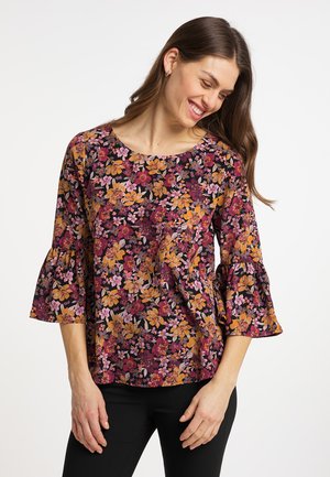 Woman smiling and looking down wearing a floral blouse with bell sleeves in shades of orange, pink, and burgundy, paired with black pants.