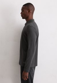 Szara koszulka polo z długim rękawem i kołnierzykiem. Miękki, teksturowany materiał o luźnym kroju, z subtelną metką z logo przy dolnym brzegu.