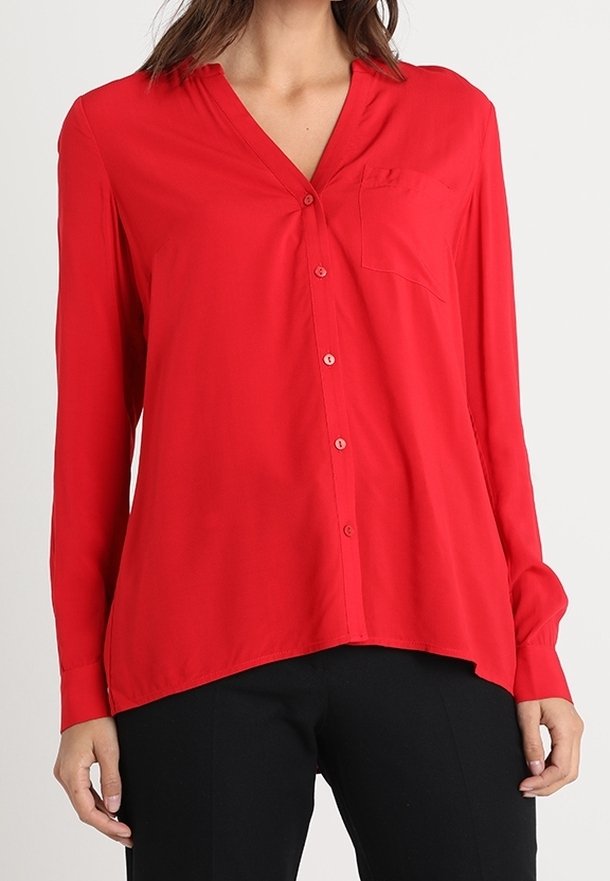 Blouse rouge à manches longues, boutonnée, avec col en V et poche poitrine, portée avec un pantalon noir.