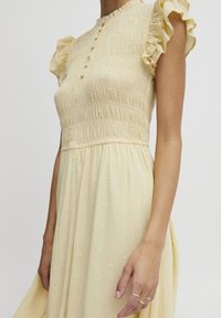 Hellgelbes Kleid mit gerafftem Oberteil, Rüschensäumen und strukturierten Punkten. Mit Knopfleiste und fließendem Rock.