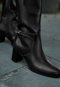 Botte cheville en cuir noir avec bout pointu, talon bloc, et bride courbée à la cheville ornée d'un accent métallique. Texture lisse avec des coutures discrètes.