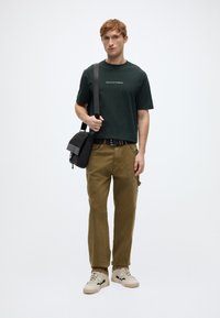 Σκούρο πράσινο κοντομάνικο μπλουζάκι με λευκό κείμενο, χακί παντελόνι, μπεζ αθλητικά παπούτσια και μαύρη τσάντα ώμου με ριγωτό λουρί.