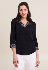 Top blu navy con scollo a V e maniche a tre quarti; presenta un colletto e polsini a contrasto con un motivo decorativo. La texture del tessuto sembra morbida e lavorata a maglia.