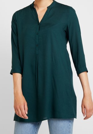 Femme portant une tunique à manches longues vert foncé avec un col mandarin et une patte de boutonnage, assortie à un jean bleu clair.