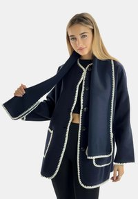 Chaqueta de mezcla de lana azul marino con ribetes y botones blancos con borde en forma de ondas, que cuenta con un cuello tipo chal y bolsillos delanteros. Bufanda ajustable incorporada.