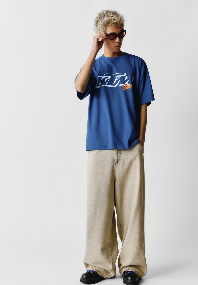 Modré tričko s bílým a oranžovým grafickým logem, spárované s volnými béžovými kalhotami a černou obuví. Model na sobě má tmavé sluneční brýle.
