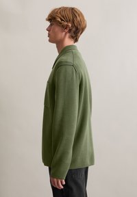 Zielony, długi rękaw, dzianinowy sweter z kieszenią na piersi, z kołnierzykiem i luźnym krojem, noszony na ciemnych dżinsach.