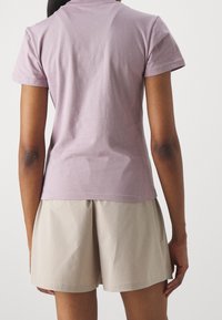 Person med en ljuslila kortärmad T-shirt och beiga veckade shorts, sedd bakifrån mot en neutral bakgrund.