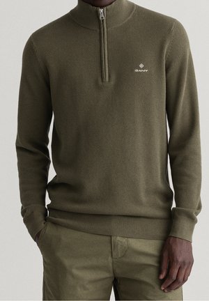 Mężczyzna ubrany w oliwkowo zielony, teksturowany sweter na zamek do połowy szyi z logo GANT oraz dopasowane oliwkowo zielone spodnie, ręce w kieszeniach.