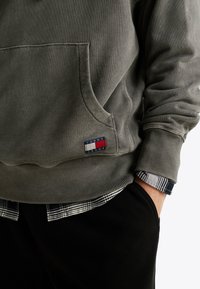 Grauer Baumwollhoodie mit einer vorderen Tasche, gerippten Bündchen und Logopatch. Darunter wird der Saum eines schwarz-weißen karierten Hemdes sichtbar.