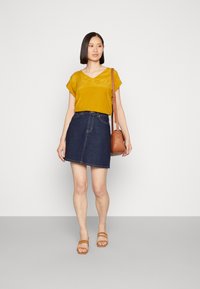 Top amarelo com decote em V e mangas roladas, combinado com uma saia em forma de A de jeans escuro. Uma bolsa castanha de ombro e sandálias bege completam o visual.