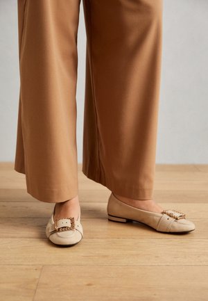Pieds portant des ballerines beige avec un détail de boucle à chaîne en or, associés à un pantalon large court couleur camel sur un sol en bois.