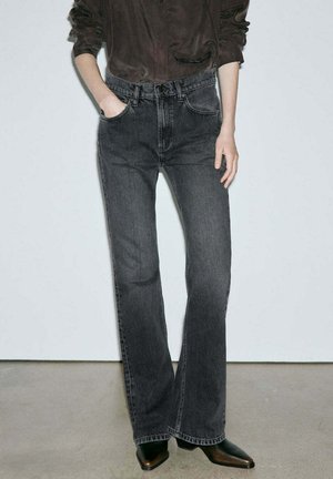Personne portant un jean bootcut noir taille haute, un haut marron foncé à manches longues et des chaussures noires à bout pointu, debout avec une main dans la poche.