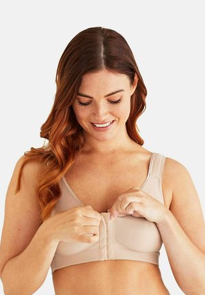 Vrouw die een beige beha met voorhaak sluit, glimlacht en naar beneden kijkt, met lang golvend rood haar tegen een effen lichte achtergrond.