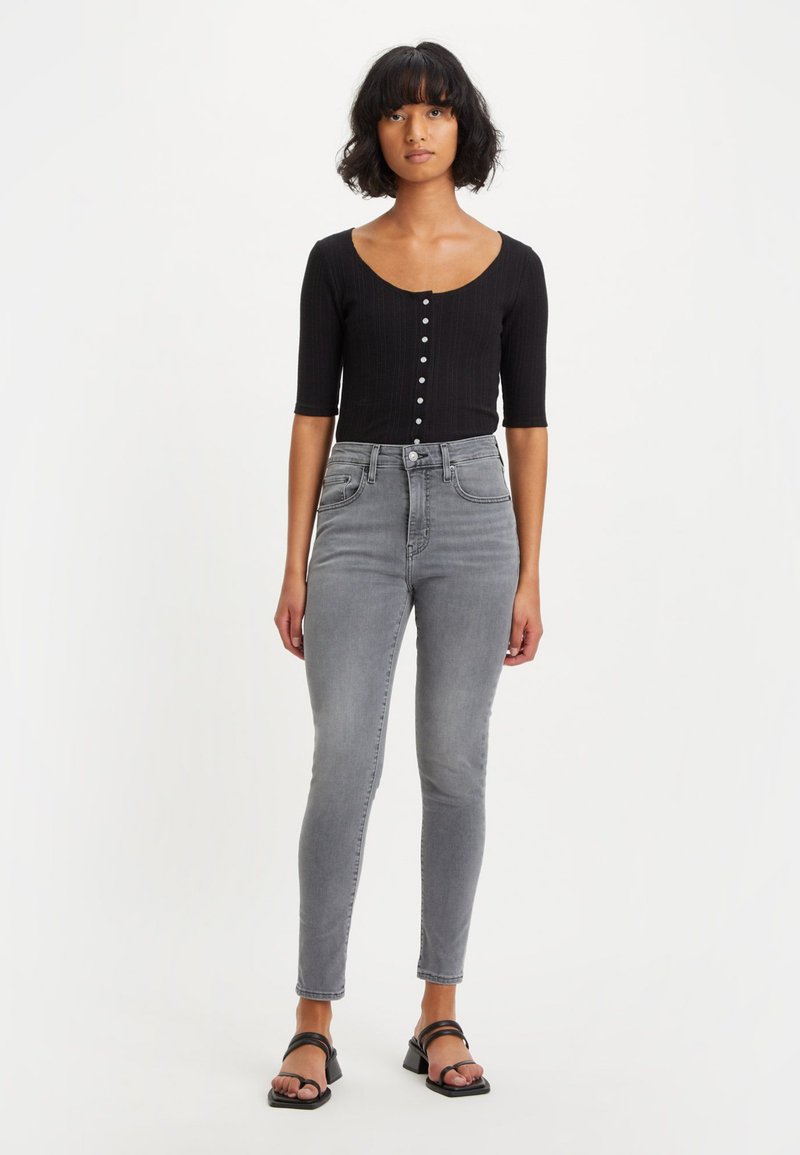 Schwarzes, geripptes Oberteil mit kurzen Ärmeln und Knopfverschluss vorne, kombiniert mit grauen, hochtaillierten Skinny Jeans und schwarzen Sandalen.
