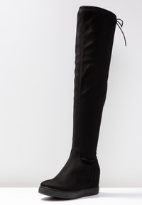 Botte noire montante au-dessus du genou en faux suède doux, dotée d'une semelle compensée et d'un lien décoratif en haut pour un ajustement réglable.