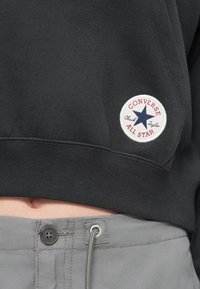 Μαύρο φούτερ με ριμπ τελείωμα, που διαθέτει στρογγυλή λευκή ετικέτα με μπλε αστέρι και κείμενο "CONVERSE ALL STAR". Γκρι ελαστική ζώνη κάτω.