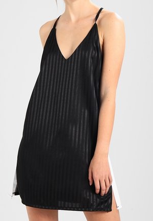 Vrouw draagt een mouwloze zwarte jurk met subtiele verticale strepen en dunne schouderbandjes, staand tegen een effen achtergrond.