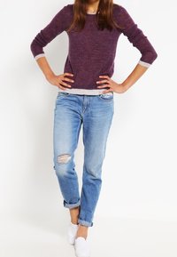 Fioletowy sweter z długimi rękawami i szarymi mankietami, w połączeniu z jasnoniebieskimi, postarzanymi dżinsami. Dopełnieniem stroju są białe buty wsuwane.