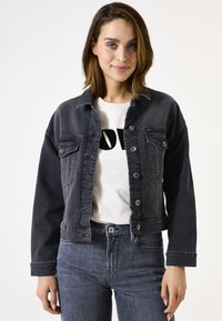 Donkerdenim jack met een getailleerd ontwerp, voorzien van knoopsluitingen, twee borstzakken en een iets cropped lengte. Gdragen over een wit shirt.