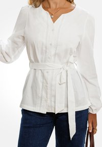 Blusa blanca de algodón con mangas largas, cierre de botones en la parte delantera, escote en forma de V y un cinturón atado en la cintura. Presenta un detalle texturizado a lo largo del placket.