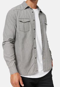 Man gekleed in een lichtgrijs overhemd met knopen over een wit T-shirt en zwarte broek, staande tegen een witte achtergrond.